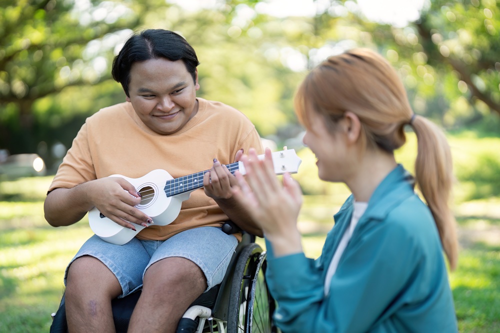 Young man in a wheelchair playing ukulele while a friend claps in admiration